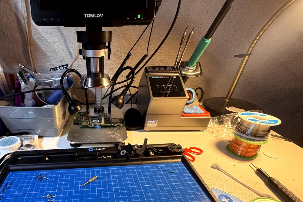 HW Repair's micro-soldering bench: a laptop motherboard positioned under a digital microscope, beside a temperature-controlled soldering station, solder and magnet wire spools, and an iFixit work mat.