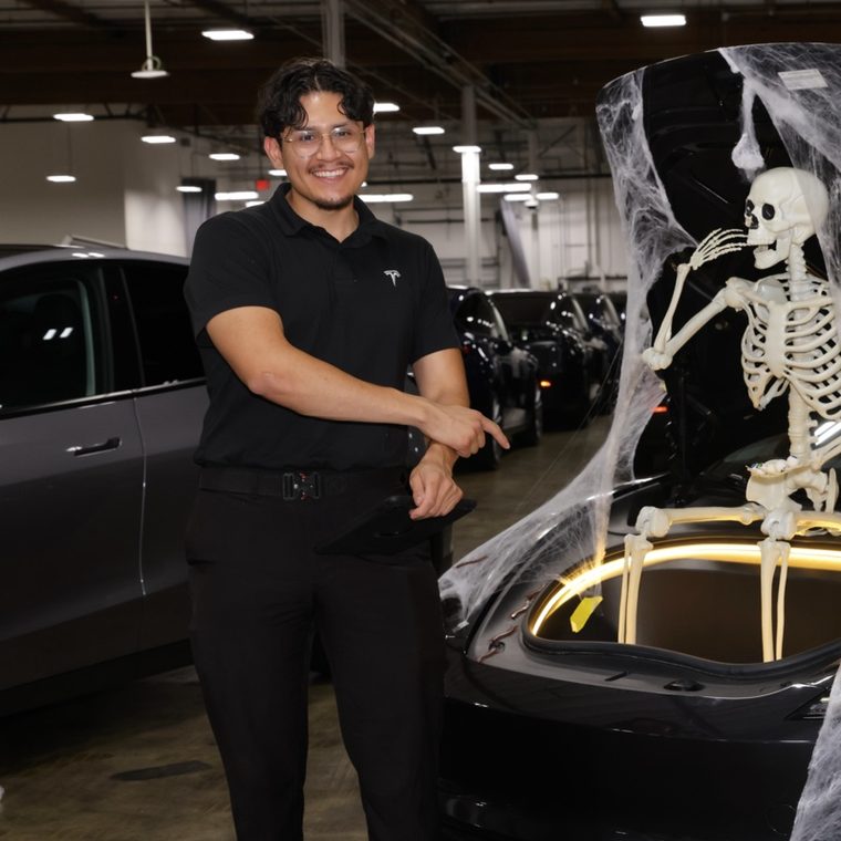 Bryan of HW Repair, smiling in a Tesla service center next to a Tesla with its frunk open and decorated with a skeleton and cobwebs for Halloween.
