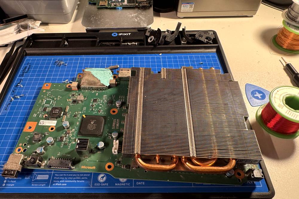 An Xbox One motherboard removed from its case on an iFixit mat, with fresh thermal paste applied and the copper heat-pipe cooler alongside it.