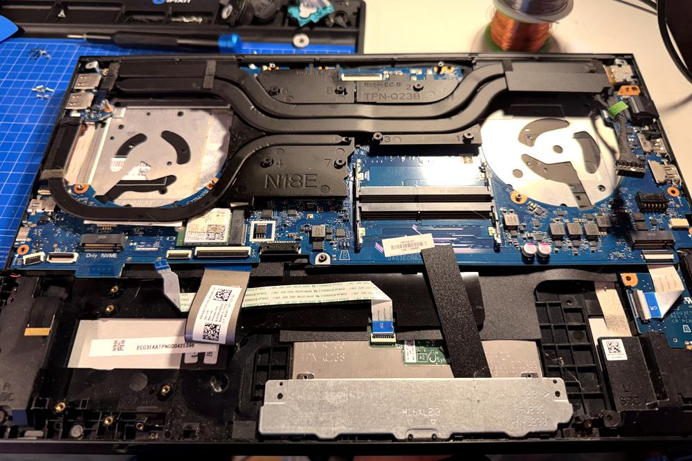 A Dell laptop opened on the bench with the bottom cover removed, showing the cooling fan, copper heat pipes, RAM slots and Wi-Fi card.